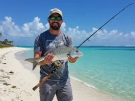 Angler holding a fish and a Florida fishing license on a sunny beach with turquoise waters and palm trees.