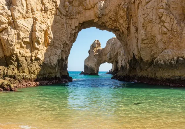 The iconic Land's End arch in Cabo San Lucas under bright sun, with clear turquoise water revealing the subtle outline of a roosterfish near the rocks.