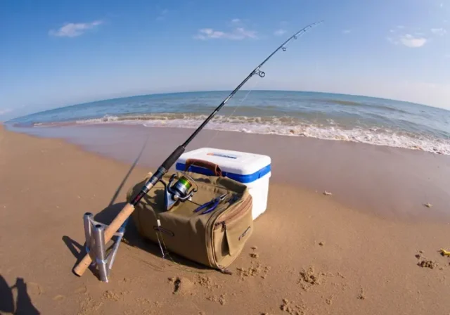 Essential must-have surf fishing gear including rod, reel, tackle bag, and cooler arranged on a beach.