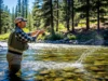 The Trout Lure Matrix: Best Lures for Every Condition A full-body shot of a man in waders standing in a clear river, casting a fishing lure on a sunny day.