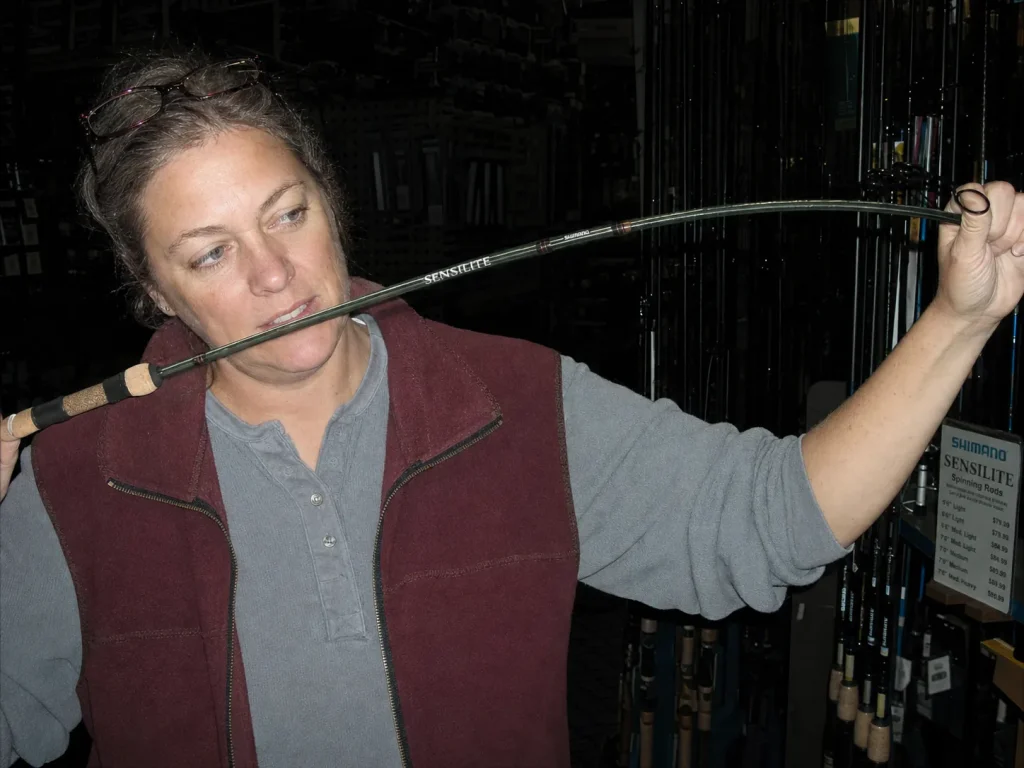 Shopper flexing a spinning rod in a tackle shop aisle under flash, judging rod feel before buying
