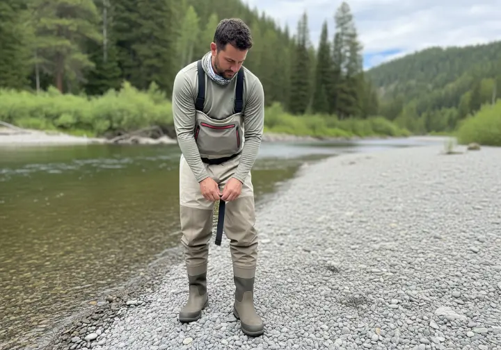 A man in fly fishing waders stands on a riverbank and adjusts his wading boot, contemplating his gear.
