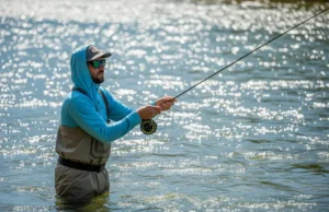 Best Fishing Sunglasses for Your Water Type: A Data-Dive A full-body shot of an angler wearing sunglasses and a sun hoodie while fly fishing in a sunny, clear river with significant glare on the water.