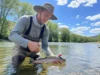 The Best Fishing Pliers: A Field-Tested Buyer’s Guide An angler in waders stands in a shallow river, using a pair of fishing pliers to carefully unhook a trout.