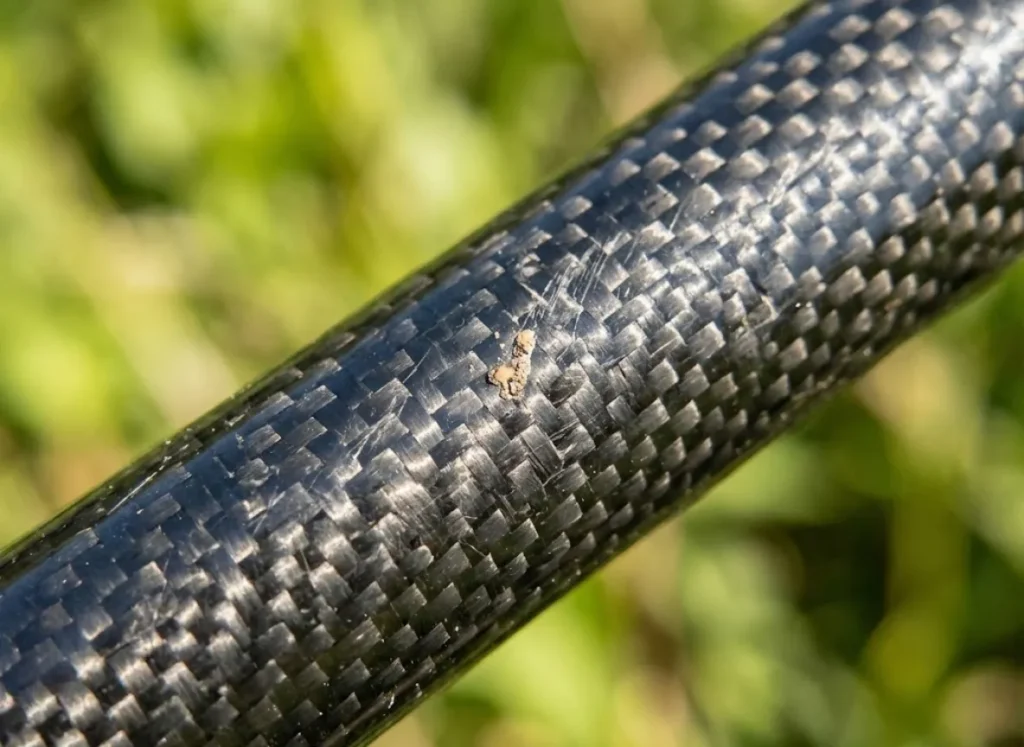 Macro photography showing the woven carbon fiber texture of a fishing rod blank illuminated by natural sunlight.