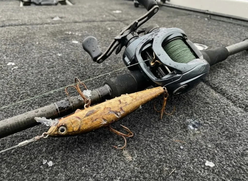 Heavy duty baitcasting rod and reel with a scarred wooden jerkbait resting on a boat deck.