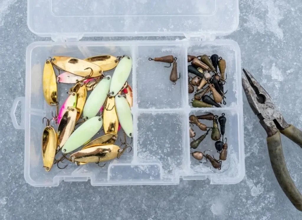 A macro shot of an ice fishing tackle box on the ice containing both gold spoons for stained water and natural jigs for clear water.
