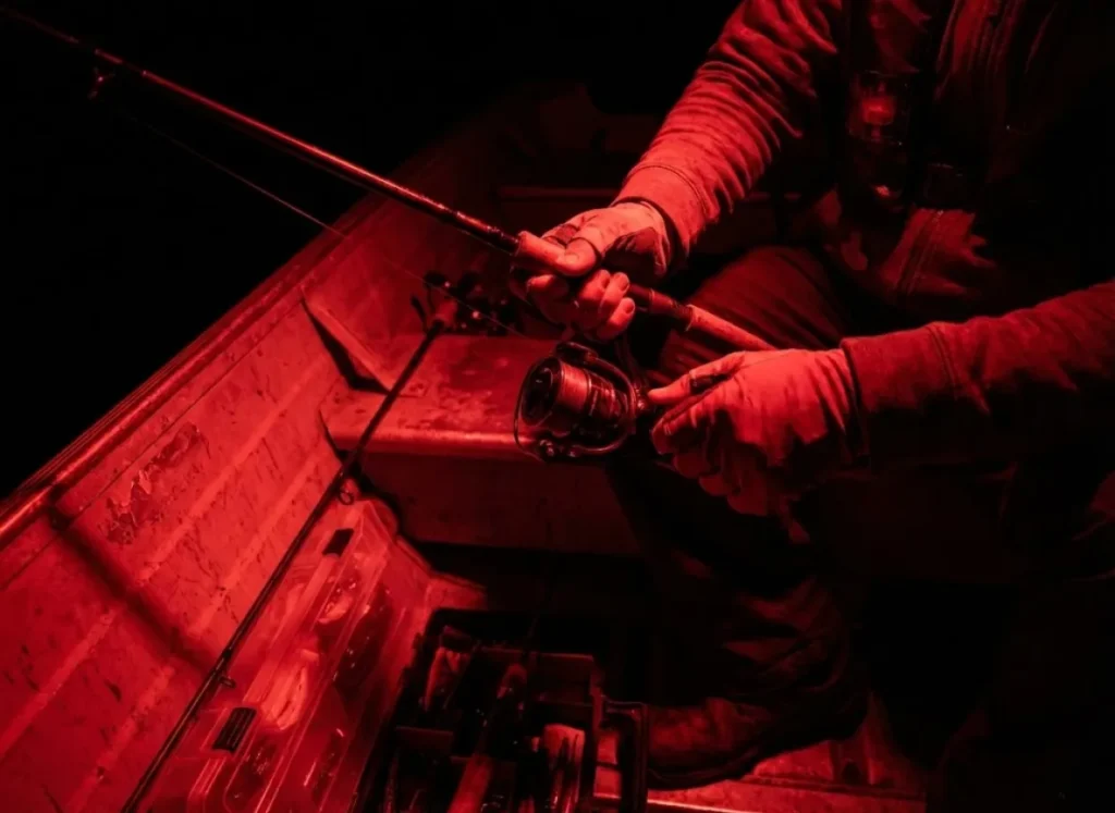 Angler adjusting a fishing reel inside a boat illuminated solely by tactical red LED lights.