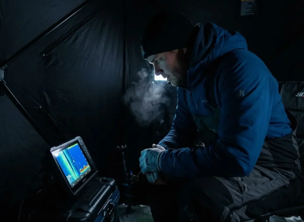 An angler inside a dark ice tent staring intently at a sonar screen while fishing for trophy Walleye.