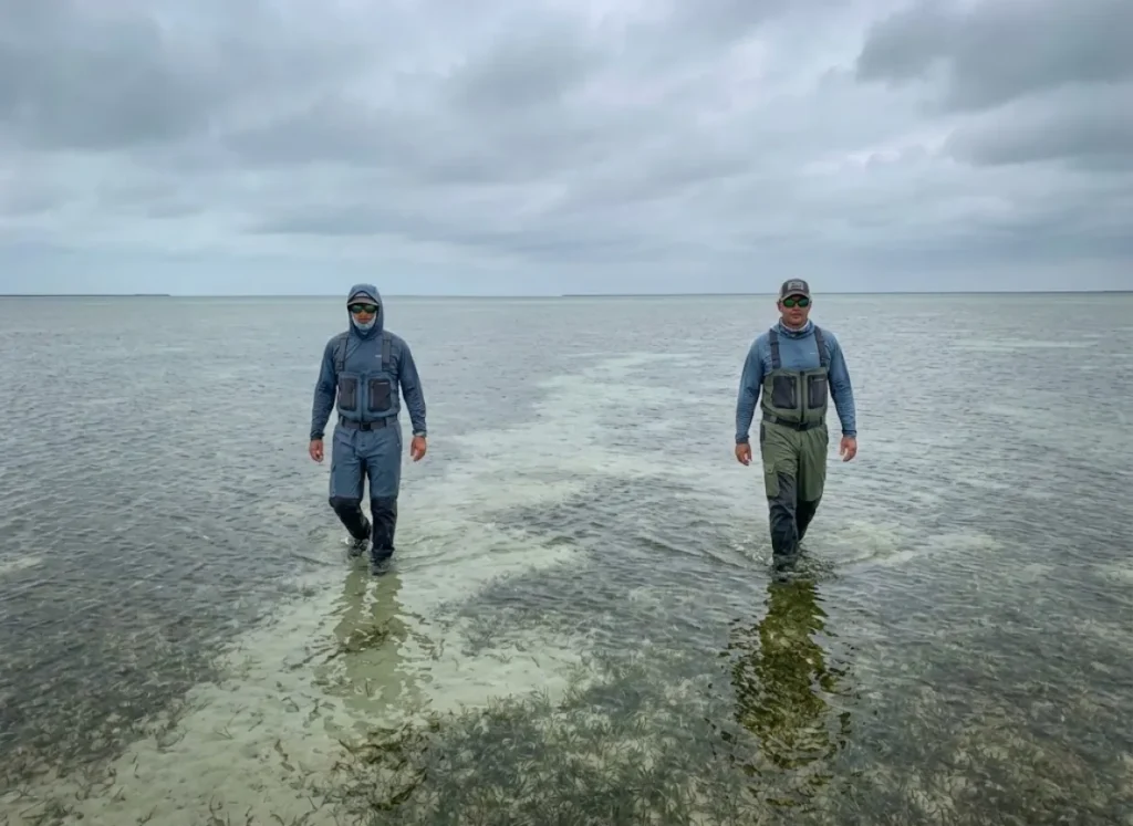Two anglers wading knee-deep across a vast, shallow seagrass flat under a cloudy sky.