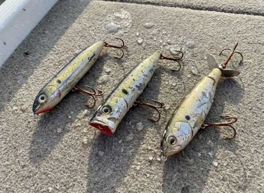 Three types of topwater fishing lures—a walker, a popper, and a prop bait—lying on a boat deck showing signs of heavy use.