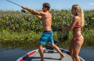 Fishing Heavy Cover: A System for Gear, Lures, and Strategy An attractive couple in their late 20s fishing for bass from a boat, casting towards thick aquatic vegetation on a sunny day.