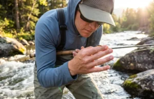 Aquatic Insects for Fishing: ID, Life Cycles & Flies A fly fisherman examining a mayfly on his finger while standing in a rushing river during sunset.