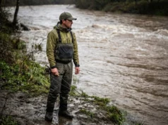 Fishing After Rain River: The Muddy Water Protocol An angler standing on a muddy riverbank looking at swollen, brown river water under an overcast sky.