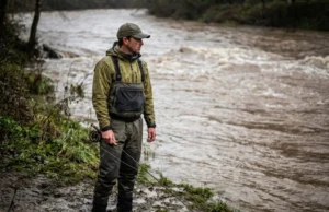 Fishing After Rain River: The Muddy Water Protocol An angler standing on a muddy riverbank looking at swollen, brown river water under an overcast sky.