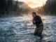 Fishing in Current: The Ultimate Water Reading Guide Angler wading in a fast-moving river at dawn, bracing against the current while holding a bent fishing rod.