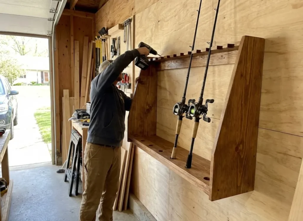 A builder securing a stained wooden vertical fishing rod rack to a garage wall with a cordless drill.
