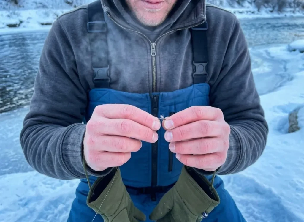 Close-up of an angler tying a tiny midge fly onto fishing line in cold winter conditions.