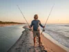 Gear Selection Guide: Saltwater vs Freshwater Durability A male angler stands on a beach between a calm river and the ocean, holding both a saltwater and a freshwater fishing rod, contemplating his choice.