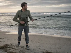 Best Surf Fishing Rods That Actually Reach Fish Angler on an open beach at dawn holding a surf fishing rod aimed at the trough