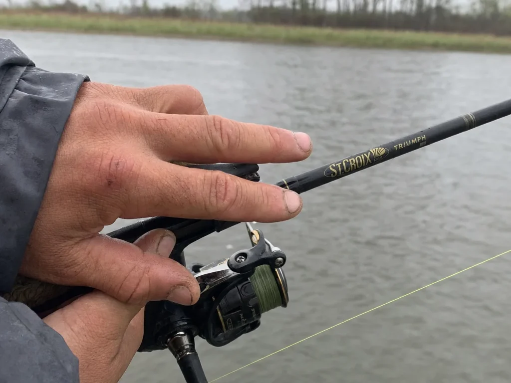 Angler's fingertips resting on a graphite rod blank feeling for a bottom bite over grey water