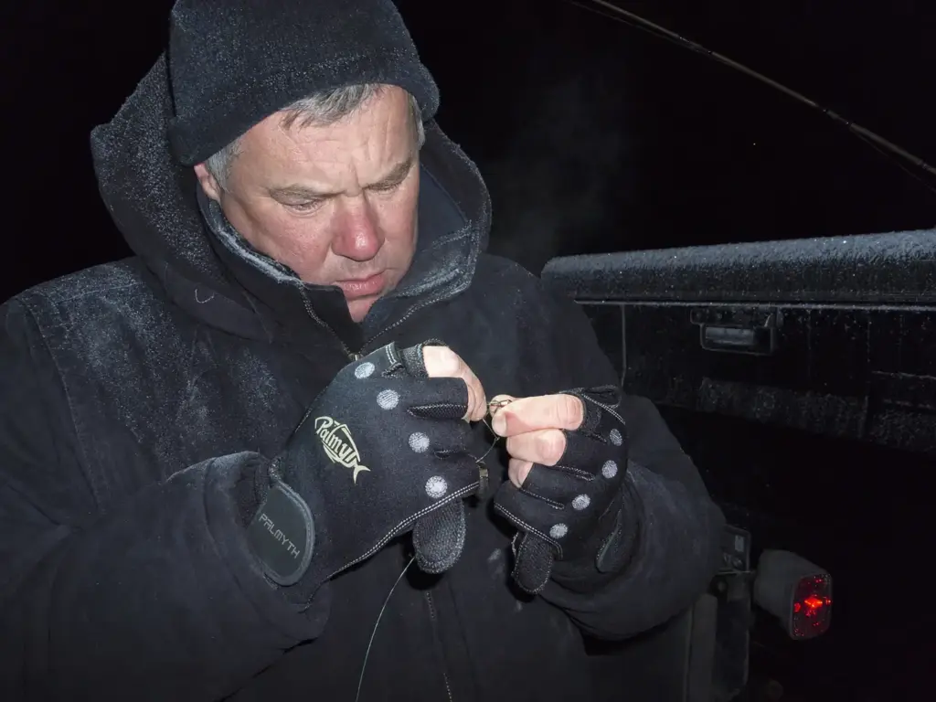 Angler in neoprene cold-weather fishing gloves struggling to tie a knot at a dark frosty ramp