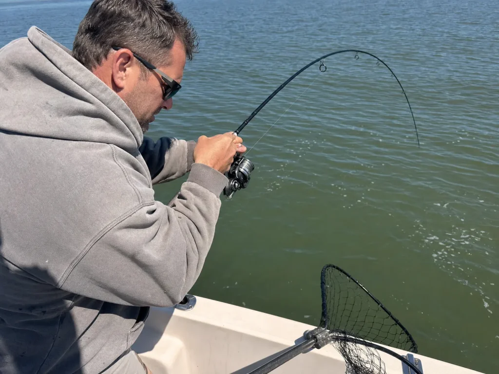 An angler fighting a fish with a deeply bent spinning rod on open gray water