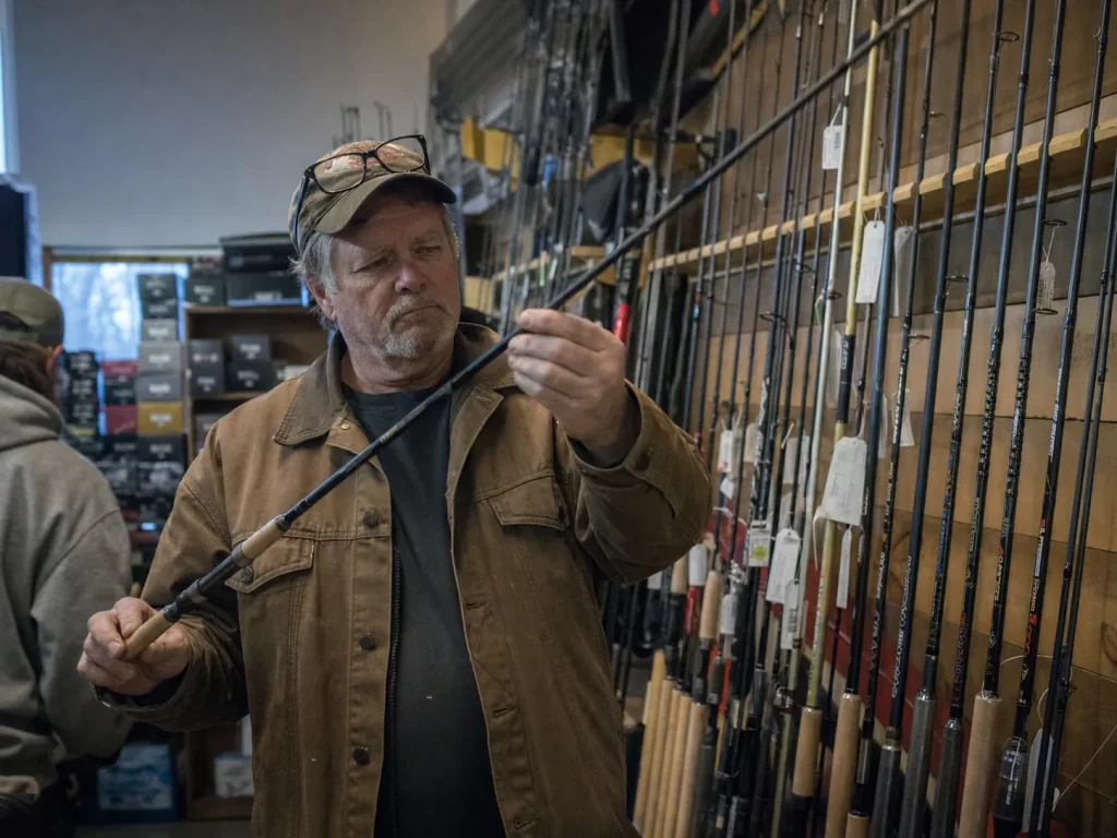 Angler holding a graphite rod off a tackle shop rack, weighing whether the upgrade is worth it