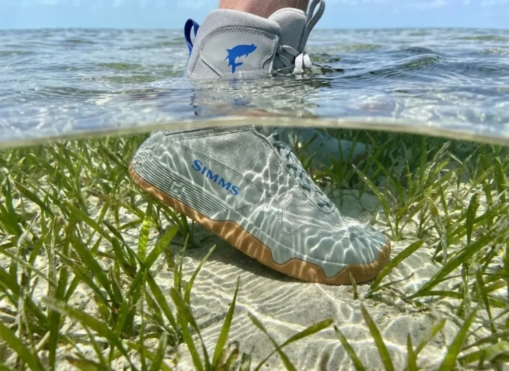 Underwater split-shot of a wading boot stepping on a sand patch amidst turtle grass, highlighting the benthic composition of the flats.