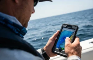 Best Fishing Apps: GPS, Tides & Maps Angler holding a rugged smartphone displaying bathymetric depth charts while fishing offshore.