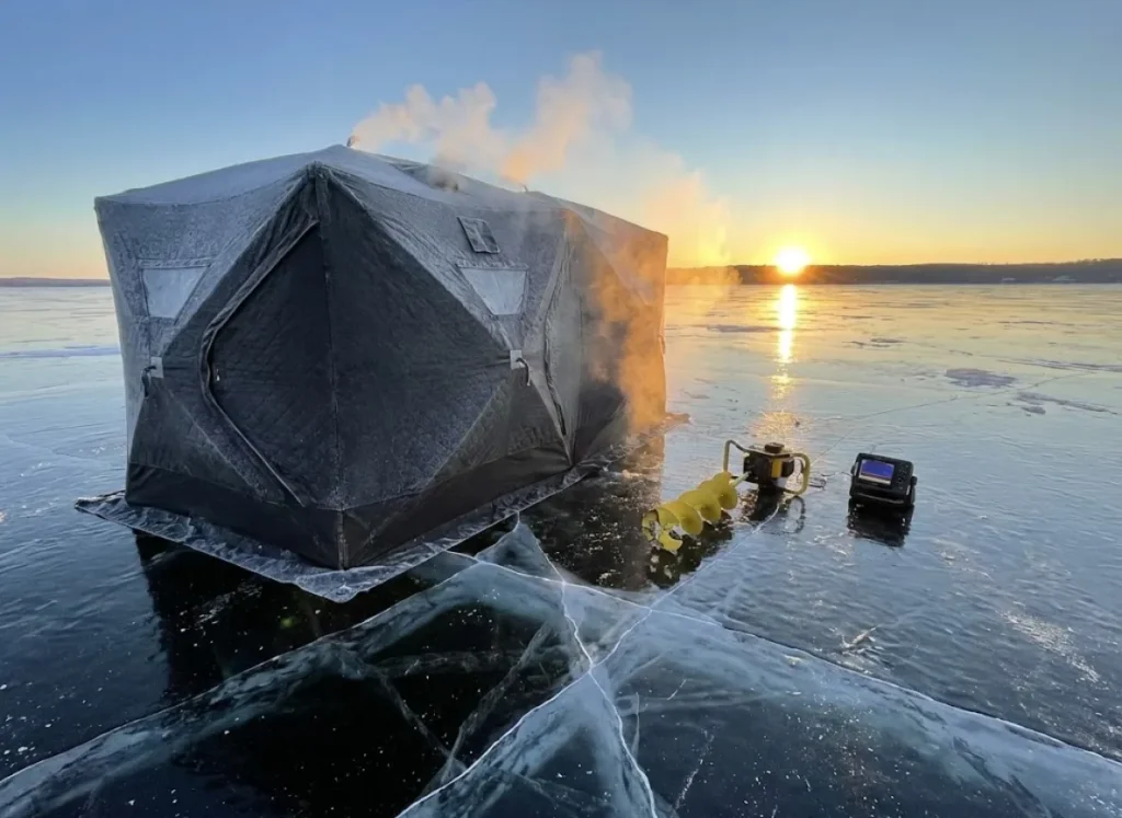 A frost-covered thermal flip-over ice shelter on black ice at sunrise with steam rising from the vent.