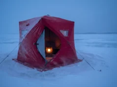 Best Ice Fishing Shelters for Wind & Cold A red insulated ice fishing shelter withstands heavy winds and blowing snow on a frozen lake at twilight.