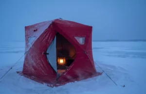 Best Ice Fishing Shelters for Wind & Cold A red insulated ice fishing shelter withstands heavy winds and blowing snow on a frozen lake at twilight.