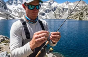 Best Swimbait Jigheads: Weedless vs Exposed Angler inspecting a swimbait jighead on a granite shoreline at a high-altitude mountain lake.
