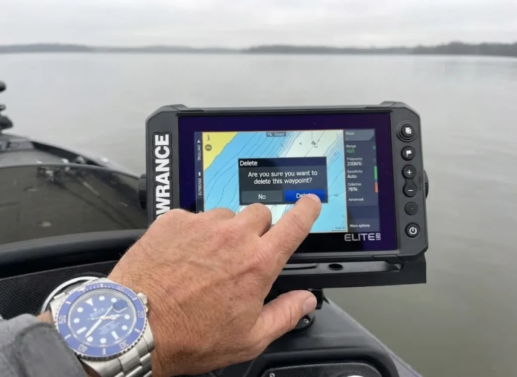 Hand wearing a dive watch pressing the delete button on a Lowrance Elite FS GPS unit to remove a fishing spot.