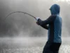 Graphite vs Glass Rods Science: The Physics of Feel An angler setting the hook on a misty lake, showing the parabolic bend of the fishing rod.