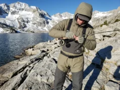 High Altitude Fishing Physics: Technical Field Manual Angler rigging fishing gear at a high-elevation alpine lake surrounded by granite peaks and snow.