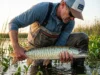 Hybrid Fish ID: Wiper & Tiger Musky Field Guide Angler in Simms waders identifying a Tiger Musky catch near reeds with St. Croix rod visible.