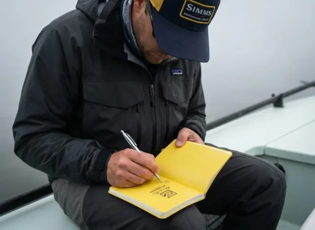 logbook-maintenance-data-audit-guide Angler writing in a Rite in the Rain notebook wearing Patagonia gear while fishing in foggy conditions.