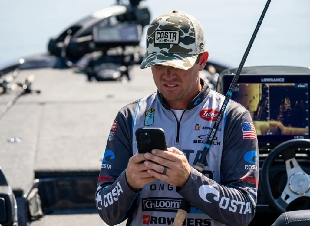 Angler holding a G. Loomis rod and checking a phone in an OtterBox case near a Lowrance fish finder.