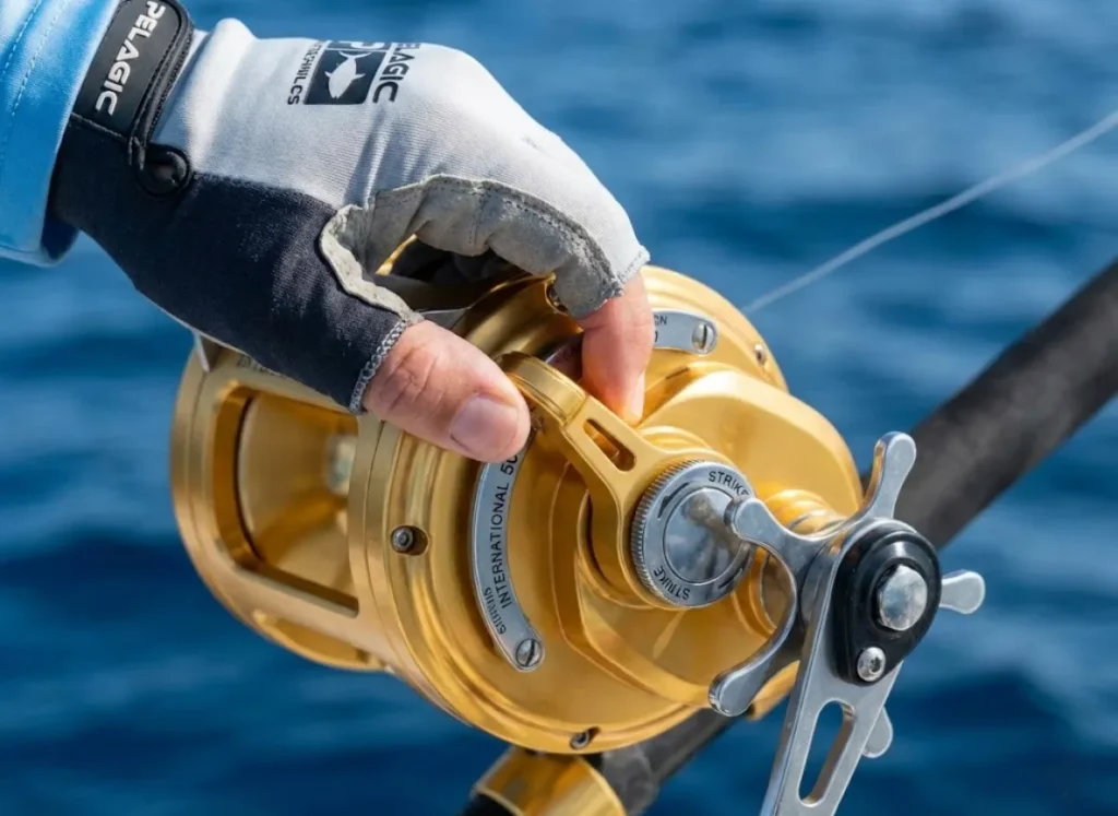 Close up of a hand in a Pelagic glove adjusting the drag lever on a gold Penn International reel against an ocean background.