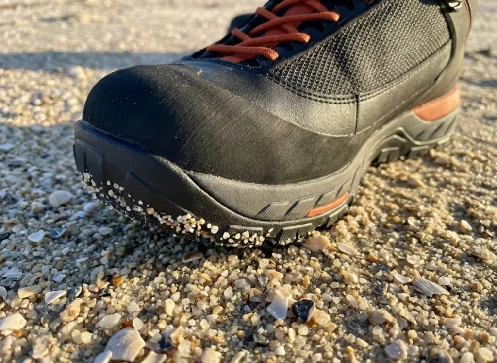 Close-up macro shot of a Simms wading boot stepping on coarse shell sand, illustrating the relationship between sediment size and beach slope.