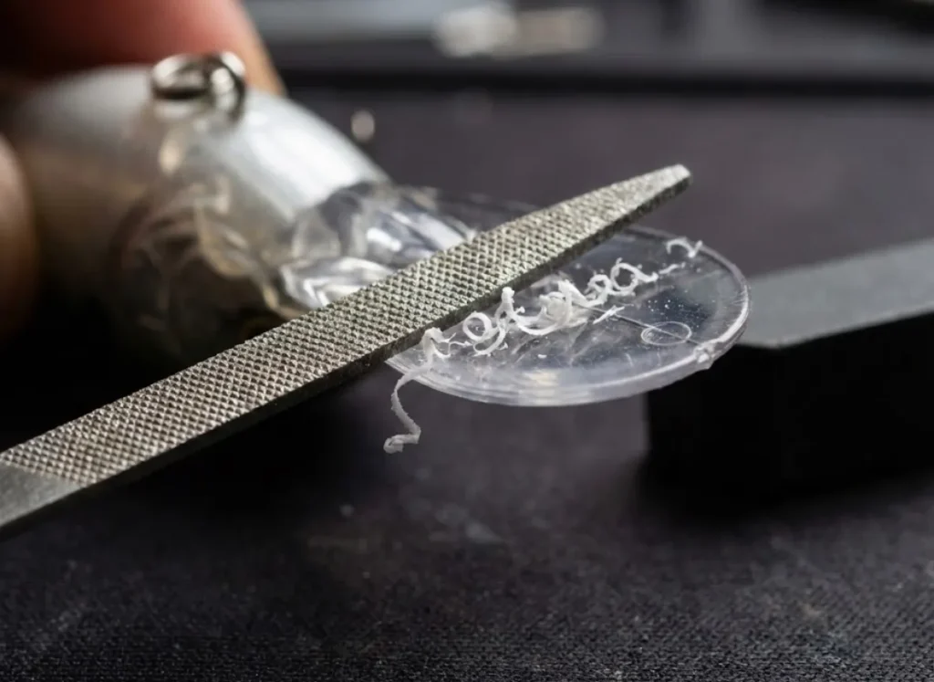 Macro close-up of a file sharpening the plastic bill of a fishing lure, showing plastic shavings and modified angles.