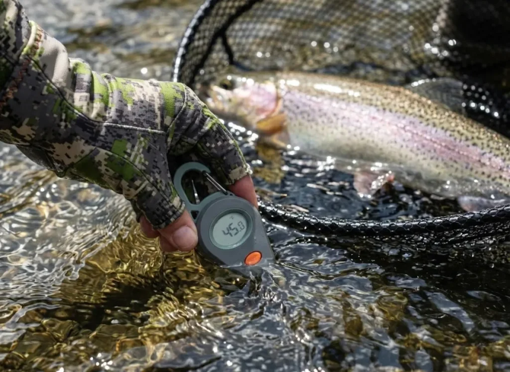 Close up of Sitka gloves holding a thermometer in water next to a trout in a Frabill net.