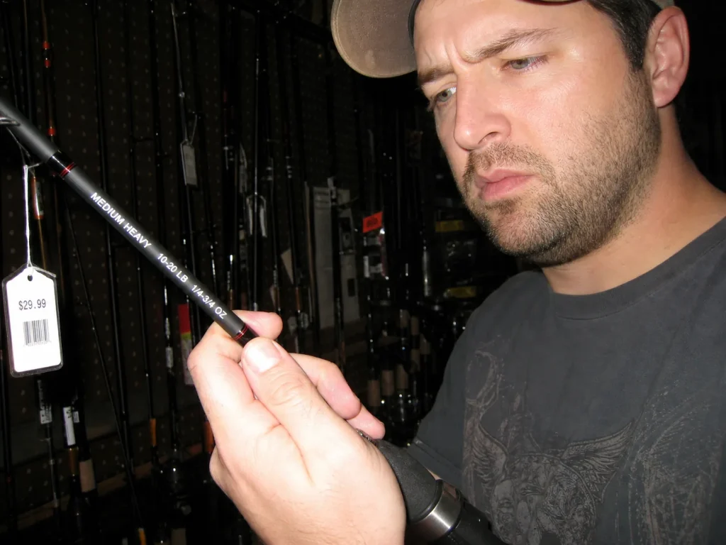 Angler in a dim tackle aisle hefting a cheap fishing rod and eyeing its optimistic printed spec label
