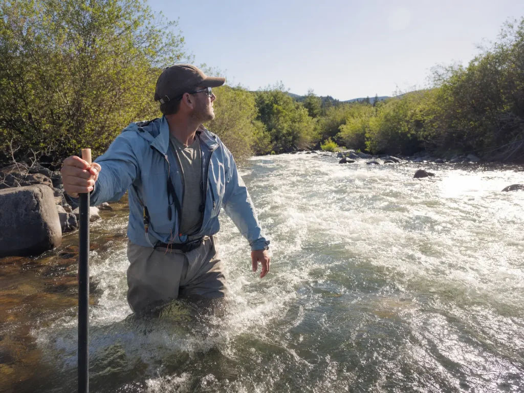 Angler pausing while wading against current on a warm day, jacket unzipped for ventilation