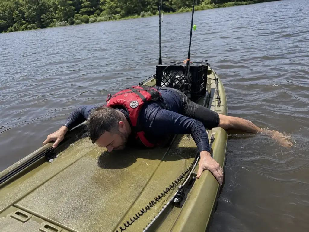 Kayak angler lying horizontally across his kayak deck mid-reentry, chest flat and legs still trailing