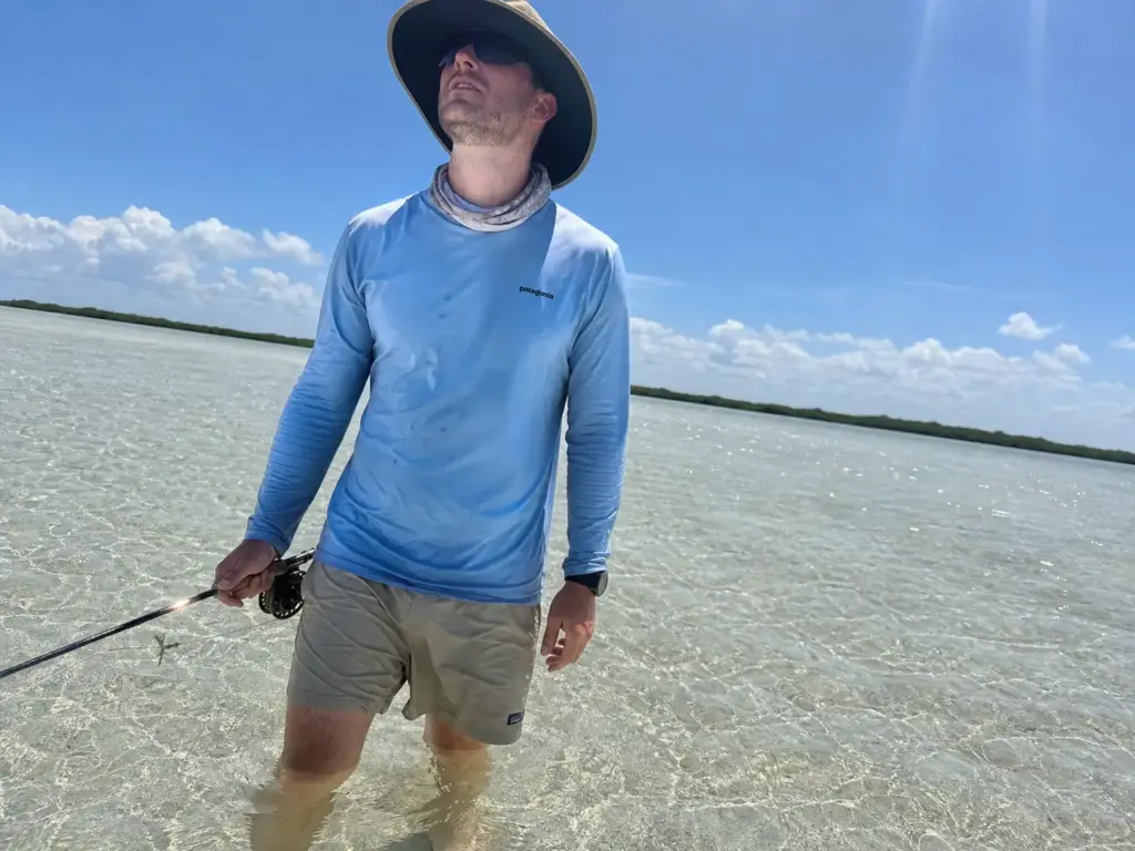 Wading angler on bright sand flats with sunlight reflecting up off shallow water under the hat brim