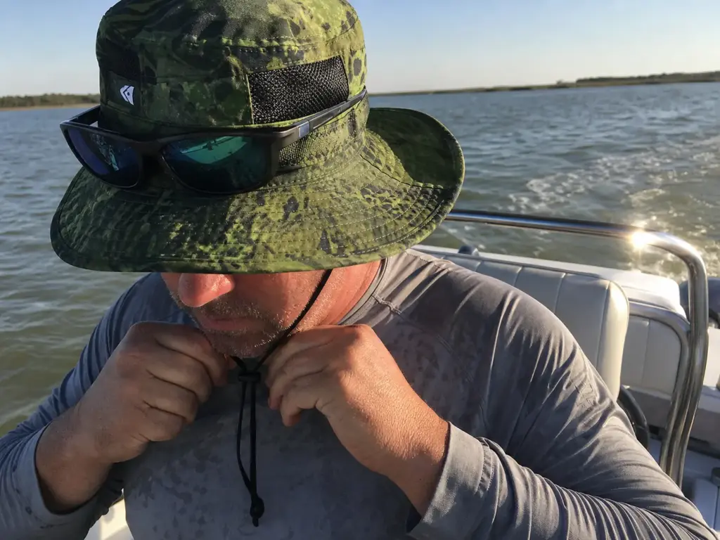 Angler cinching the chin strap on a vented KastKing boonie hat in wind on a running boat