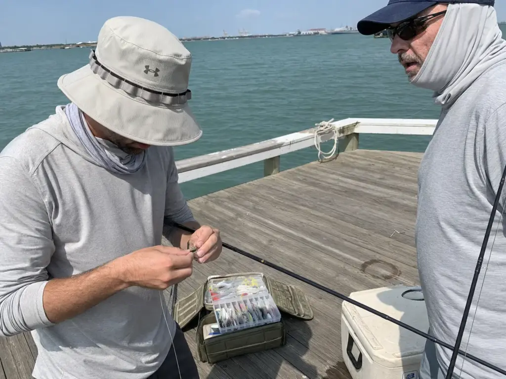 Two anglers on a dock, one in a bucket hat and one in a cap with a buff pulled up over the neck